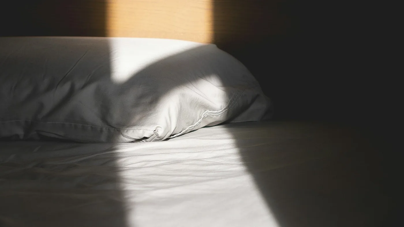 朝日が斜めに差し込む枕元。シーツのしわに光の帯が落ちている